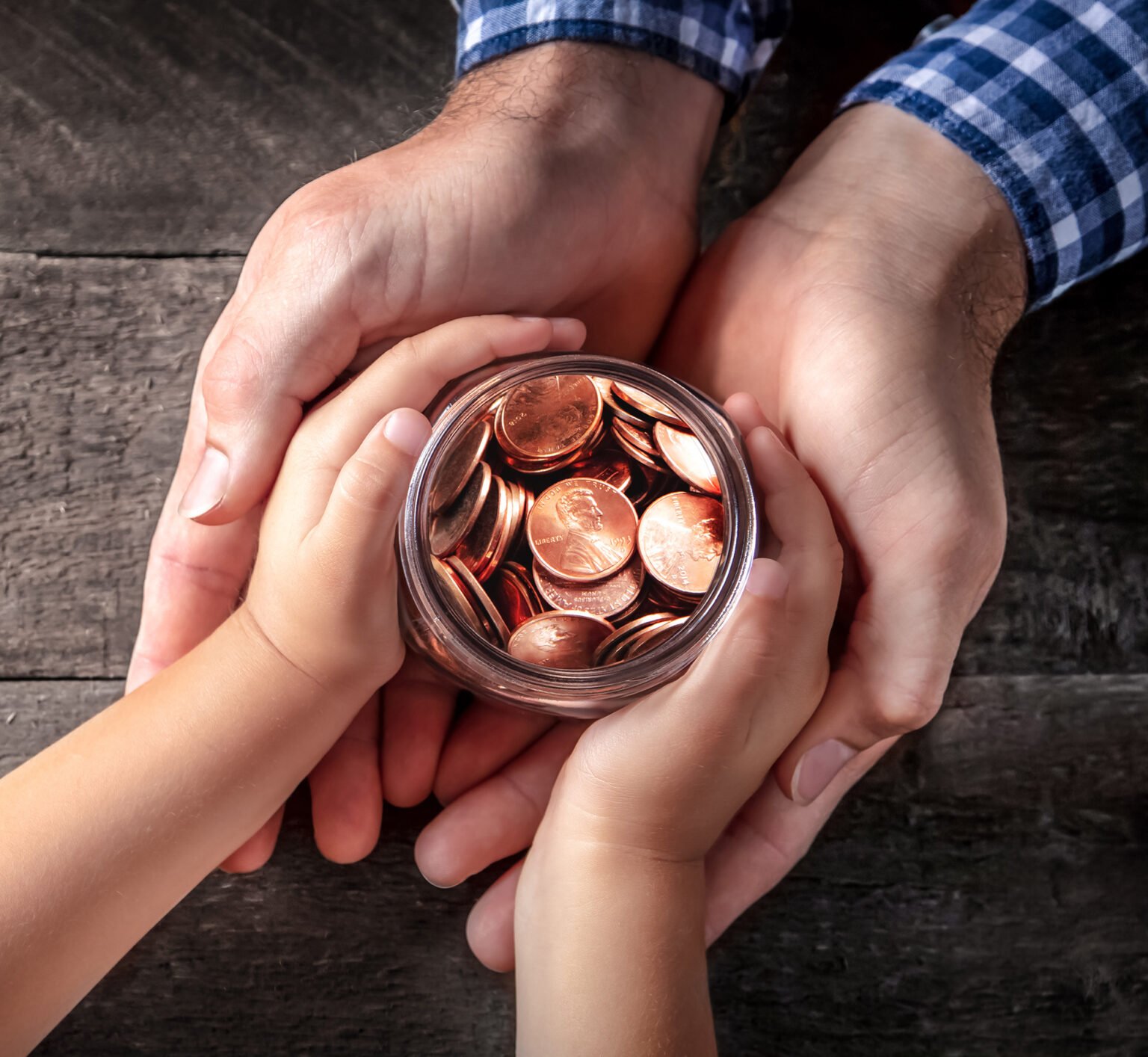 Child and parent hold a jar of coins on a wood table.